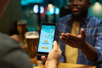 Focus on screen of smartphone held by young man ordering taxi to bar while sitting in front of his buddy explaining him something