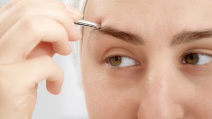 Closeup portrait of smiling young woman forming eyebrows with tweezers after having bath. Concept of beautiful female, makeup at home, skin care and domestic beauty industry.