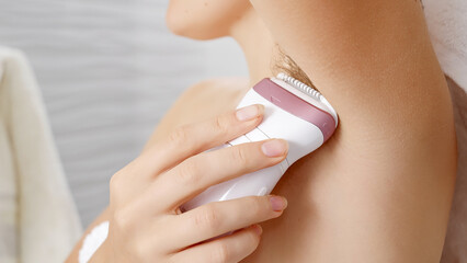 Young woman using electric epilator to remove armpit hair in bathroom