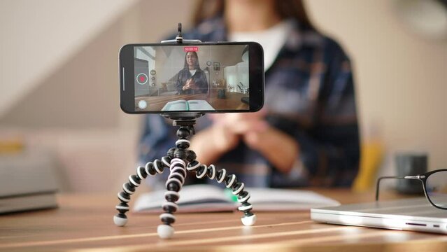 Smiling woman blogger vlogger sitting at desk recording educational video on mobile phone on tripod, sharing knowledge, streaming webinar for followers. Young female creating content for vlog, blog.