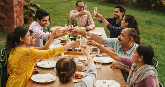 Big Indian Family And Friends Celebrating At Home, Diverse Group Of Children, Young Adults And Senior People Gathered At The Table To Have Fun Conversation. Clinking Glasses And Making Toast