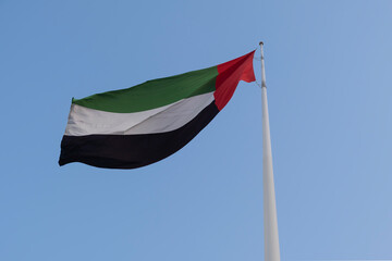 United Arab Emirates UAE flag waving on a mast against blue sky, shot from the bottom, looking up.