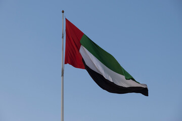 United Arab Emirates UAE flag waving on a mast against blue sky, shot from the bottom, looking up.