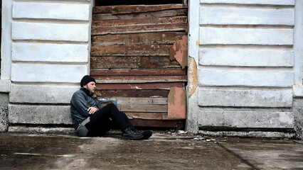 Homeless man sits outside an abandoned building. Refugee needs help. Below poverty line. Illegal immigrant. Tramp wearing in dirty clothes and hat cap. Fugitive. Consequences of drunkenness alcoholism