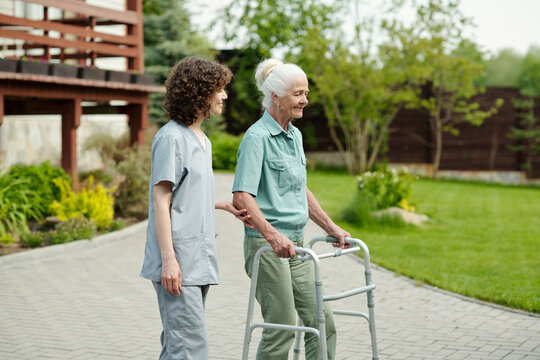 Young caregiver supporting hand of senior patient of retirement home using walker during stroll in the garden with green lawns and trees