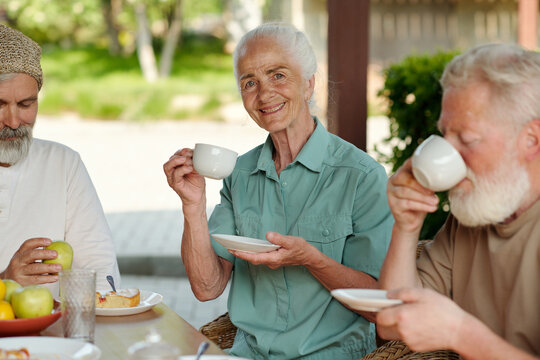 Happy senior woman with cup of tea and saucer looking at camera with smile while sitting by served table between two elderly men