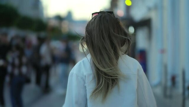 The camera from the back captures a girl walking down the street among young people with long hair and a white shirt