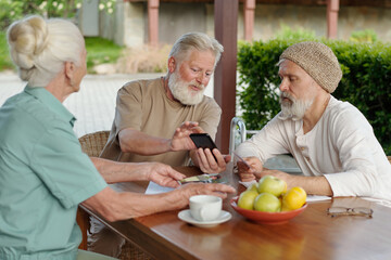 Aged bearded man showing photos of his family in smartphone to senior male and female patients of retirement home sitting next to him