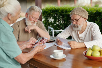 Two elderly bearded men and grey haired woman looking through photos of their family and grandchildren while sitting by table after breakfast