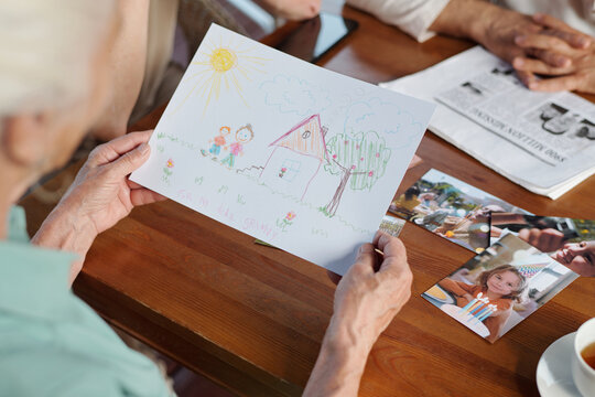 Focus on drawing of child held by elderly woman sitting by wooden table with photos of her grandchildren and looking through them