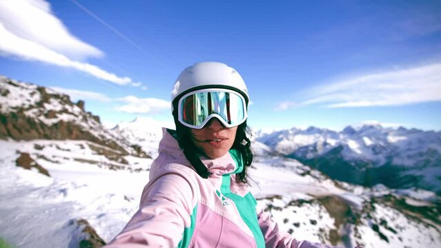 Smiling woman blogger vlogger standing on top mountain talking with camera recording video. Female skyer in protective clothing helmet goggles. Create content for blog, vlog. Winter sport resort.