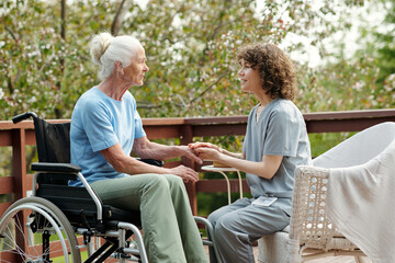 Side view of young caregiver holding hands of senior female with disability sitting in wheelchair...