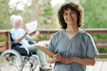 Happy young nurse or caregiver with clipboard looking at camera with smile while standing against...
