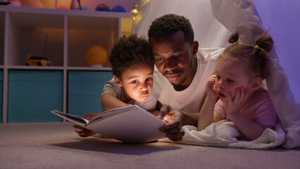 African american dad and son, caucasian daughter reading book lying in wigwam.