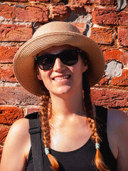Young woman straw hat portrait on red brick wall urban background. Millennial girl smiles walks on summer city street with bright sunshine. Stylish confident authentic lady in black sunglasses outdoor