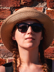 Young woman in straw hat portrait on red brick wall urban background. Millennial girl walking on summer city street with bright sunshine. Stylish confident authentic lady in black sunglasses outdoors.