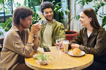 Two happy young intercultural women with glasses of latte chatting and looking at one another while sitting against guy with glass of water