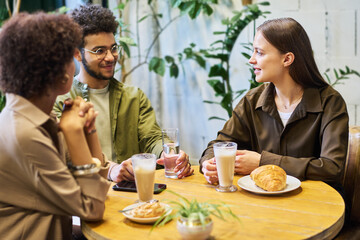 Young smiling woman with glass of latte looking at bearded guy during chat while both sitting in front of female friend in cozy cafe