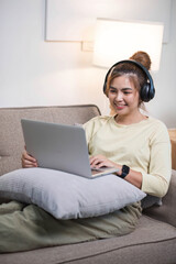 Young beautiful asian woman relaxing and listening to music using headphones, she is lying in sofa. Young woman enjoying at home using laptop.