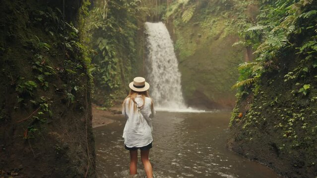 Woman tourist in straw hat taking picture recording video on camera of huge waterfall among tropical mountains on island. Travelling in wild nature admiring views. Travel tourism, vacation concept.