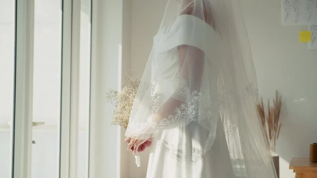 Cropped shot of bride in white wedding dress and veil standing in salon with bouquet of flowers in front of window