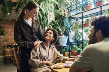 Young brunette woman looking at one of her friends with smile and keeping hands on her shoulders during communication in cafe