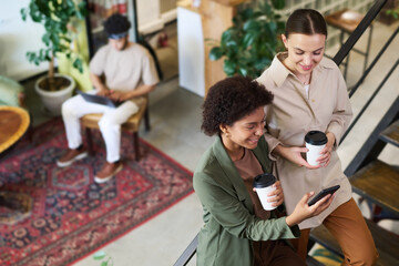 Two young smiling intercultural women in smart casualwear looking at smartphone screen during...