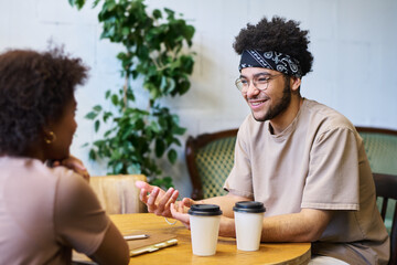 Fototapeta na wymiar Happy young multiethnic man in casualwear talking to his girlfriend or colleague while sitting by table with two cups of coffee in cafe