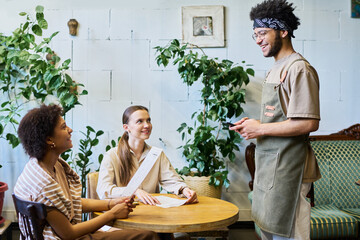 Two intercultural girls with menu looking at smiling waiter in workwear standing by their table and waiting for their orders in cozy cafe