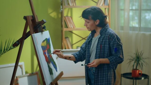 A beautiful woman with short pixie haircut is painting something on a canvas while also holding a brush in her mouth and enjoying her time doing so