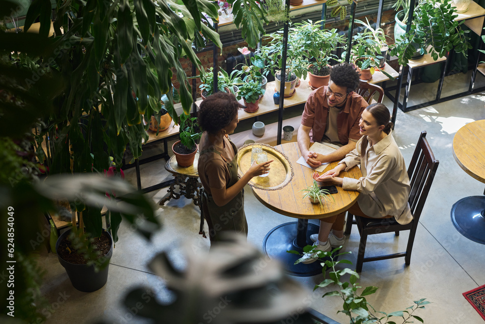 Poster high angle of young intercultural couple looking at waitress with tray standing in front of their ta