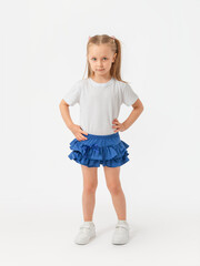 A 5-year-old girl stands tall on a white background and looks expressively at the camera in a white T-shirt, blue skirt and sneakers.