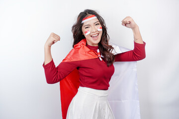 Excited Asian woman wearing a red top, flag cape and headband, showing strong gesture by lifting...