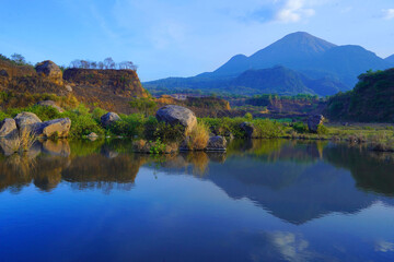 Fototapeta na wymiar A serene pond reflects the surrounding landscape of lush grass, scattered rocks, and towering trees by the breathtaking view of mount Penanggunan in Ranu Manduro, Mojokerto, East Java, Indonesia.