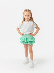 A cute 5-year-old girl in a T-shirt and skirt is standing, her hands on her waist, her leg pushed forward in sneakers on a white background. Looking into the camera