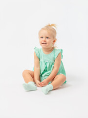 A cute 2-year-old girl is sitting looking up and smiling on a white background in socks and a green dress. Expressive look.