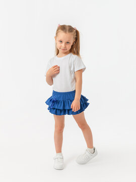 A modest shy 5-year-old girl with an expressive look in a white T-shirt, blue skirt and sneakers stands on a white background.