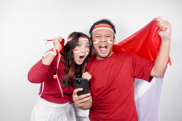 A young Asian couple with a happy successful expression watching match oh their phone while holding Indonesia's flag, isolated by white background. Indonesia's independence day concept.