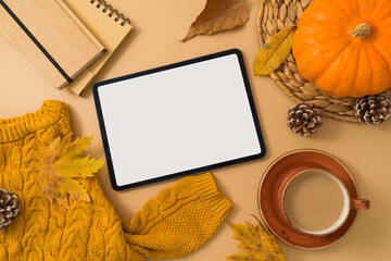 Autumn season concept with digital tablet mock up, coffee cup, warm sweater, fall leaves and pumpkin on beige background. Top view, flat lay