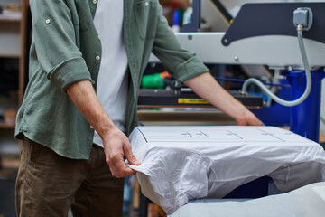 Cropped view of young craftsman holding t-shirt with marking while working with screen printing machine in print studio at background, customer-focused small business concept - obrazy, fototapety, plakaty