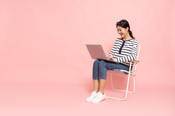 Portrait of an excited Asian woman sitting and typing keyboard on laptop computer isolated on pink...