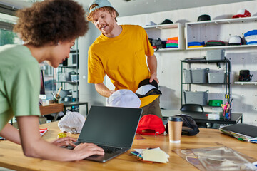 Smiling young designer holding snapbacks while talking to african american colleague using laptop near color swatches in print studio, small business resilience concept