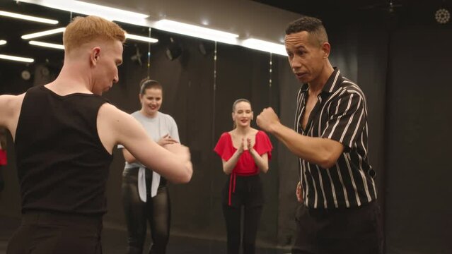 Medium slowmo of two young expressive multiracial men dancing salsa and girls clapping to them while having group Latin dance class in ballroom