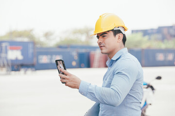 Male logistic foreman using mobile phone browsing the internet and looking at camera at container...