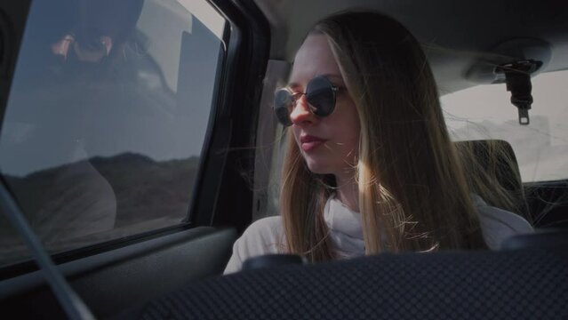 A beautiful young girl in a car in the back passenger seat looks out the window at the mountains. Enjoying the journey. Road trip and Wanderlust. Long hair moving in the wind, sunglasses.