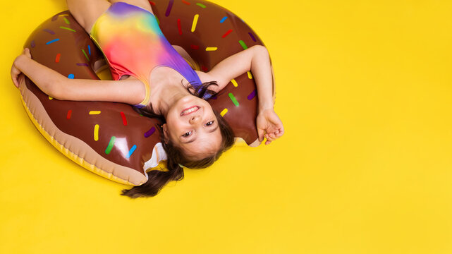 upside down girl in one-piece rainbow colored swimsuit swims in inflatable ring on yellow background with place for text