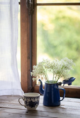 Brocca di latta di colore blu con fiori di colore bianco e tazza banca e blu su tavolo di legno di fronte ad una finestra