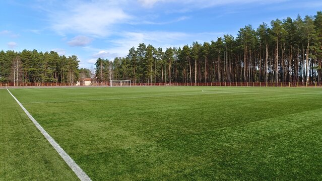 The stadium is located among pine trees and surrounded by a fence. The soccer field has an artificial surface, a goal with a net and markings. Sunny spring weather and blue sky with clouds