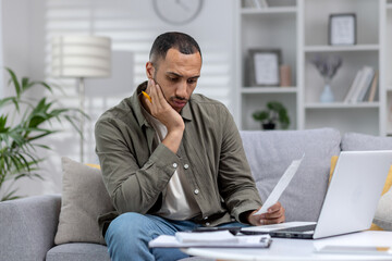 Tired and thoughtful african american man working from home on laptop with documents and bills. Overtime over the deadline - Powered by Adobe
