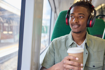 Young Man Commuting To Work Sitting  On Train Wearing Wireless Headphones With Takeaway Coffee